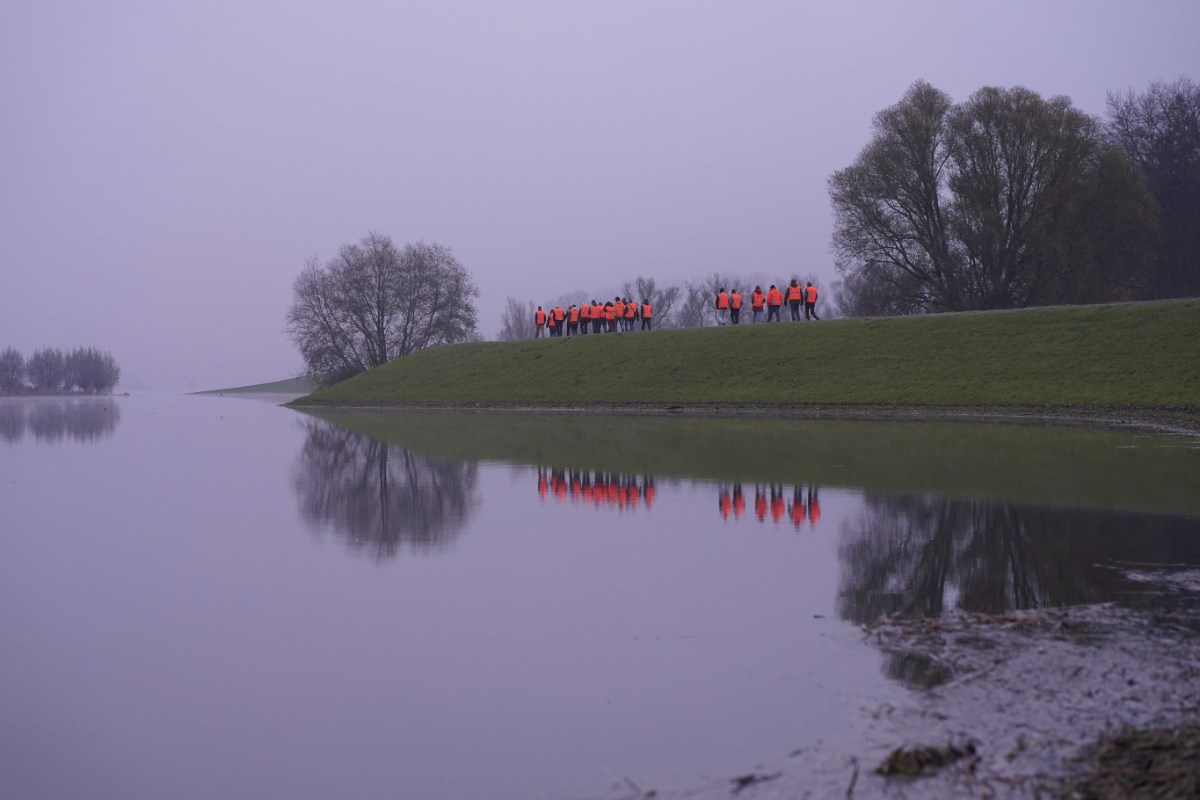 Een groep mensen in oranje hesjes loopt over de dijk. Op de voorgrond stroomt de IJssel.