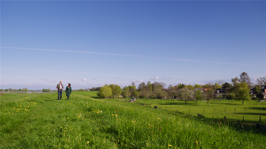 Twee personen lopen op het gras van de dijk. De lucht is strakblauw en de personen zijn in gesprek.